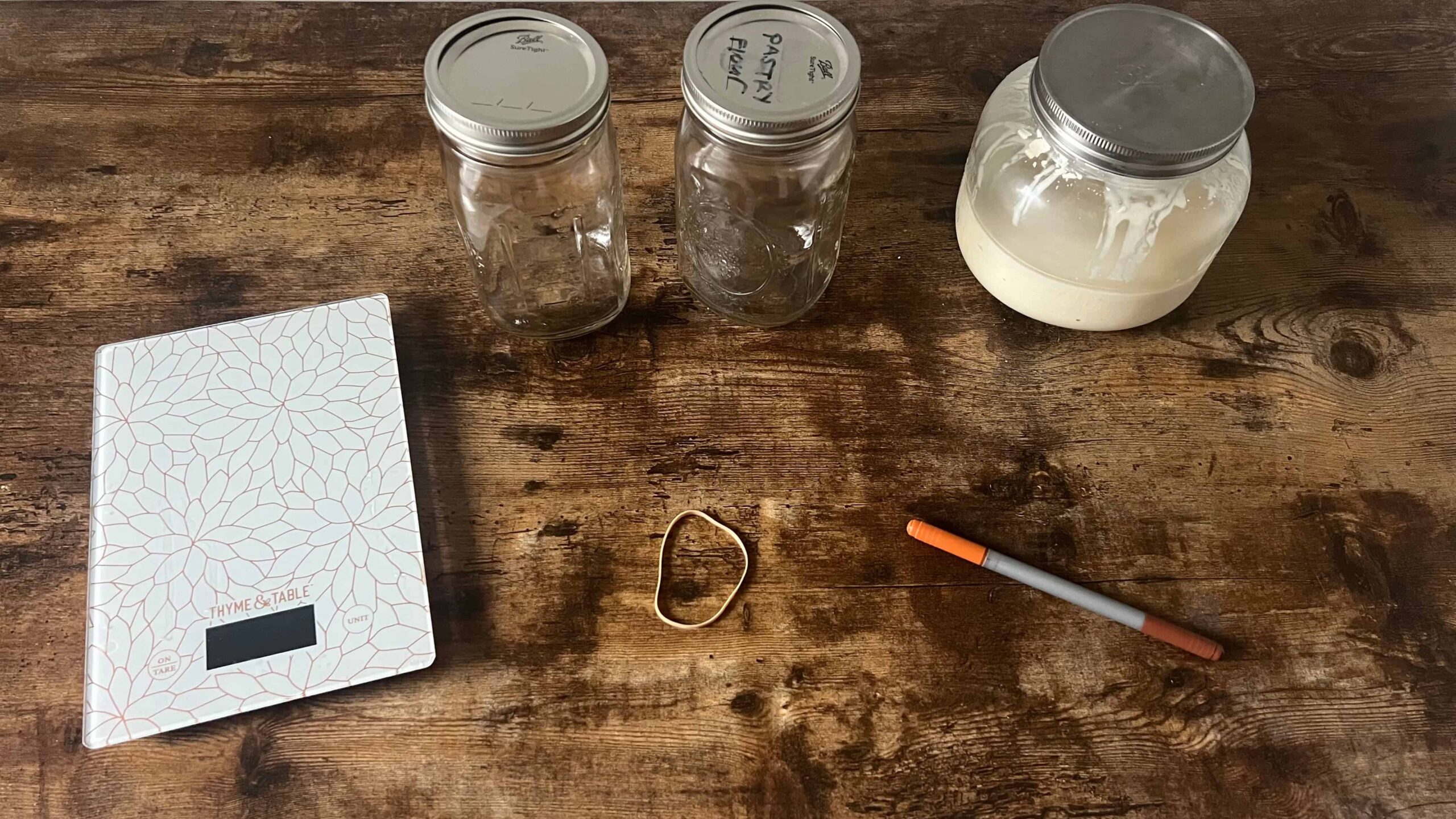 A digital kitchen scale, two empty mason jars, a mason jar with sourdough starter, a rubber band and a marker are laid on a brown table. These are the tools needed for feeding a sourdough starter. 