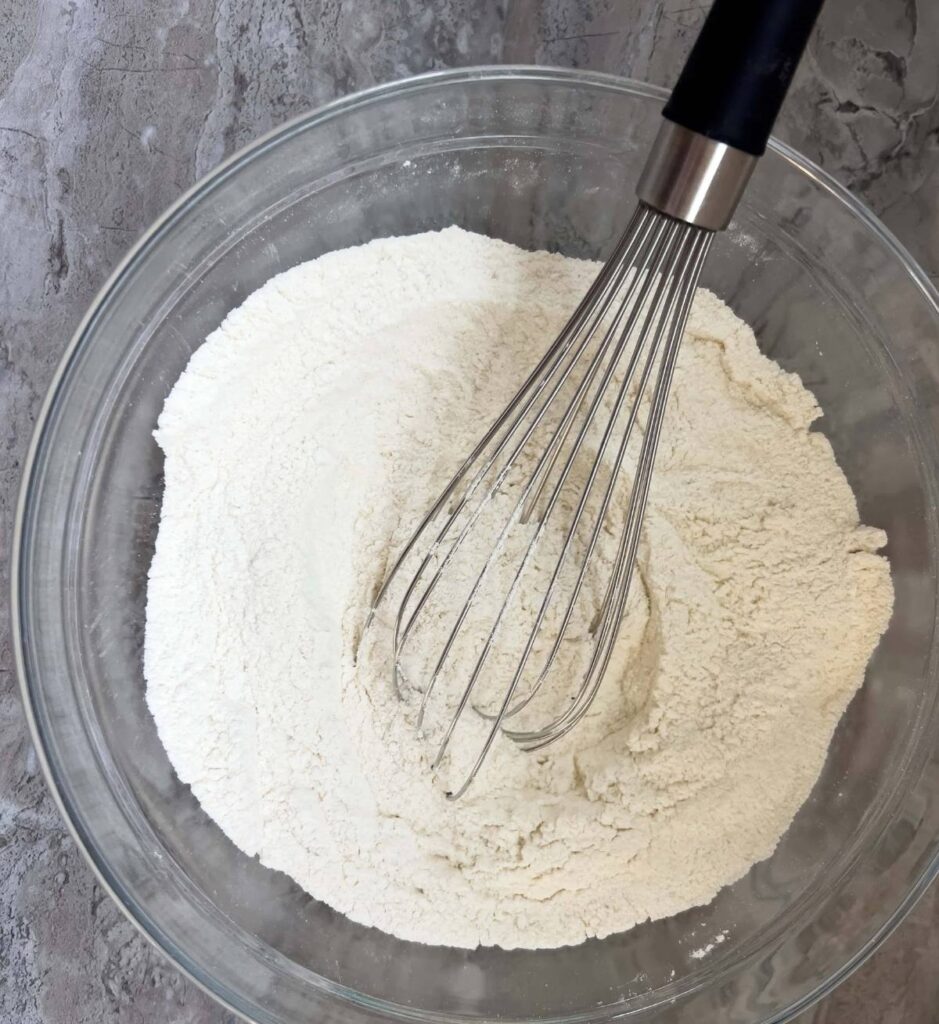 Dry ingredients including flour, sugar, baking powder and baking soda mixed in a glass bowl with a wire whisk resting in the bowl.