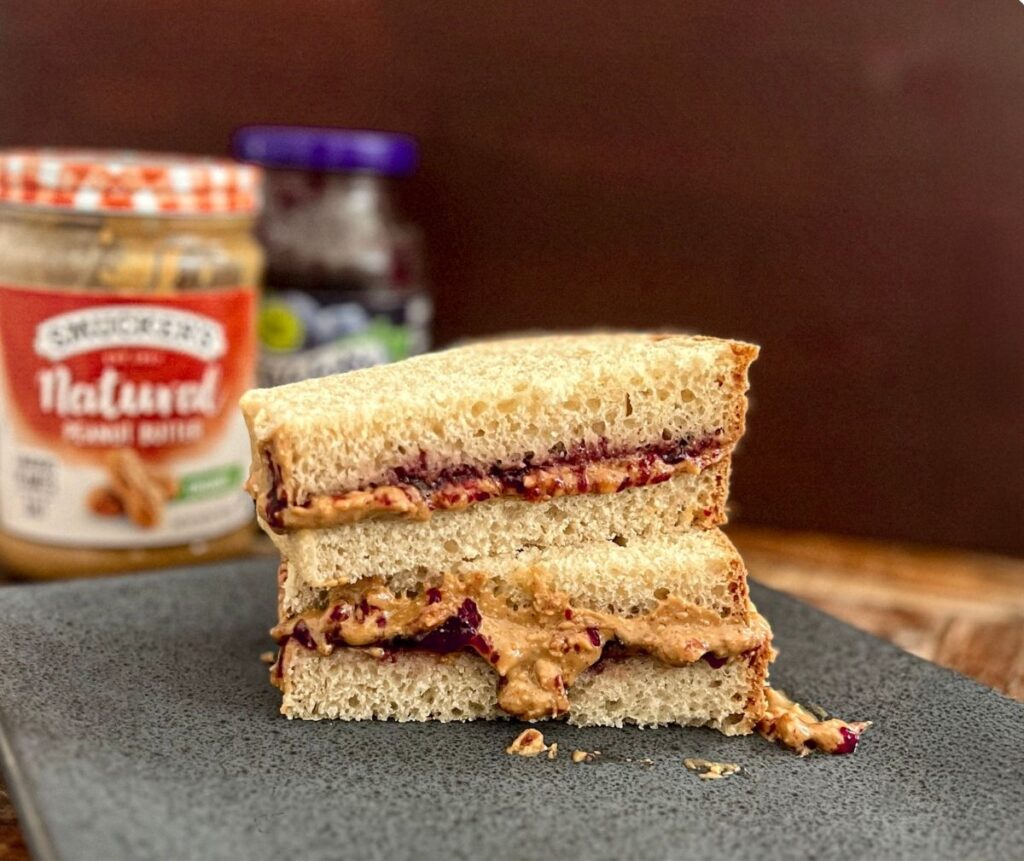 A peanut butter and jelly sandwich using honey wheat sourdough sandwich bread cut in half and stacked with a jar of peanut butter and a jar of jelly in the background.