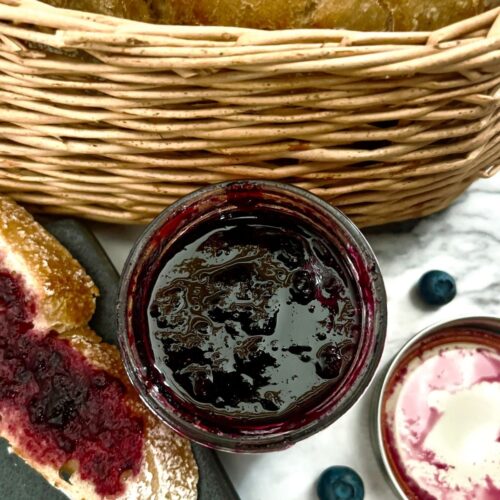A glass jar of easy blueberry bourbon preserves (no pectin) next to a bread basket and sliced bread.