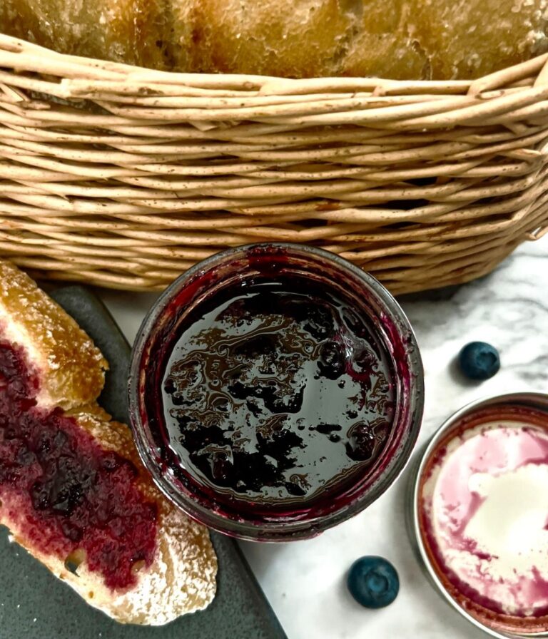 A glass jar of easy blueberry bourbon preserves (no pectin) next to a bread basket and sliced bread.