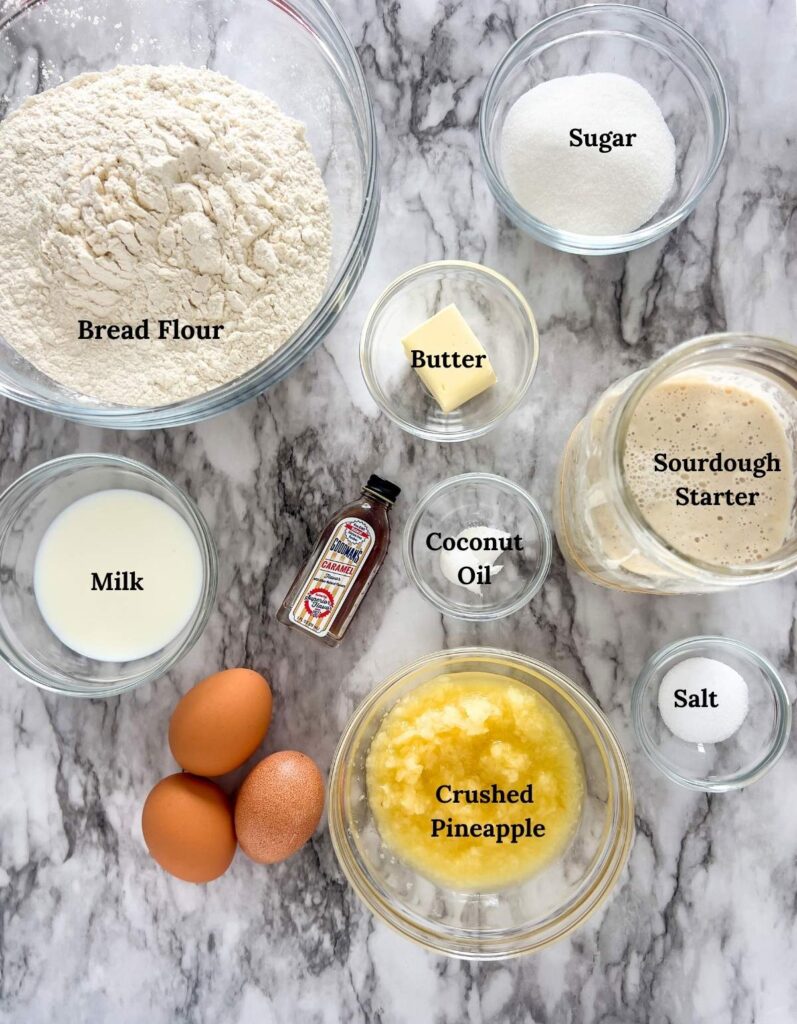 Ingredients for sourdough Hawaiian rolls in glass bowls on a marble countertop.