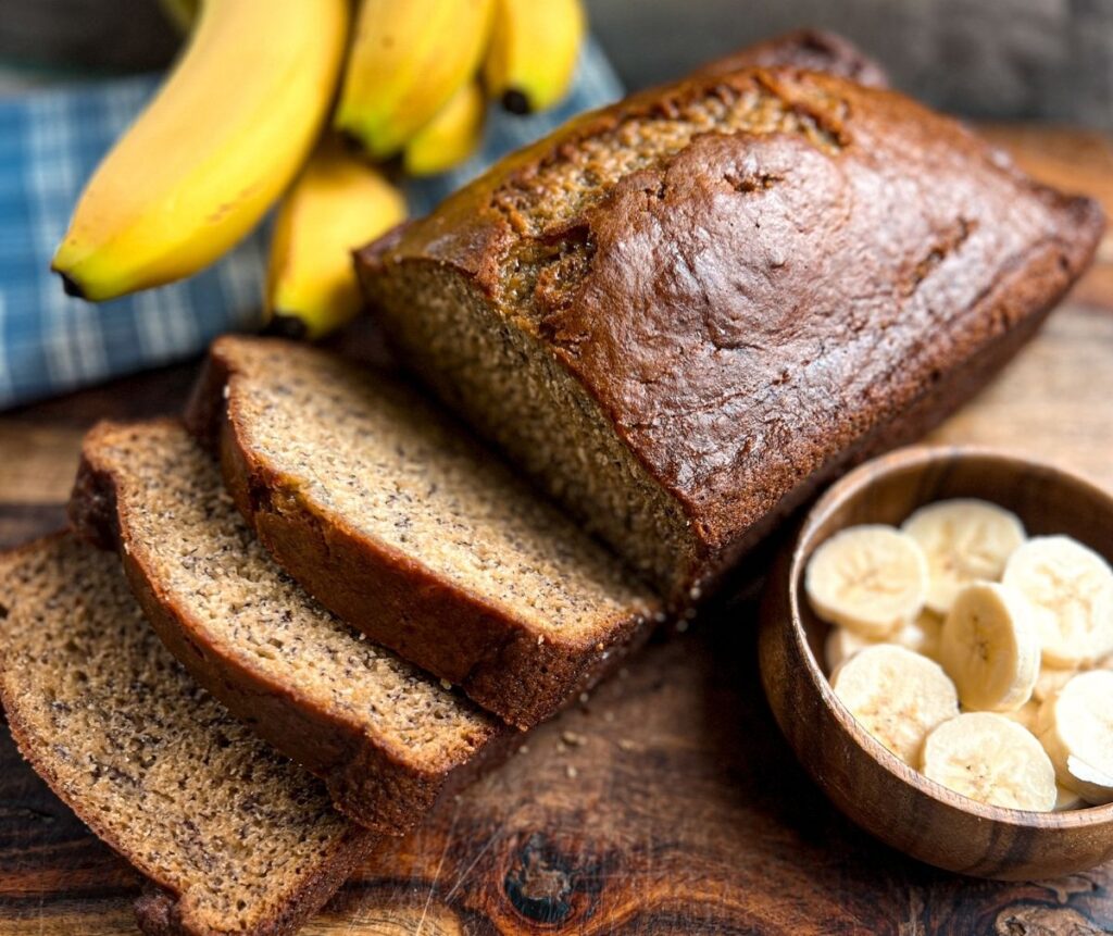 Slices of no oil banana bread next to a bowl of banana slices and whole bananas in the background.