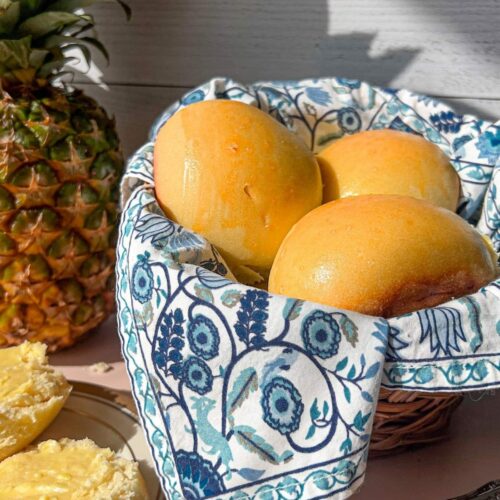 Sourdough Hawaiian rolls resting in a blue cloth lined basket with a pineapple in the background.