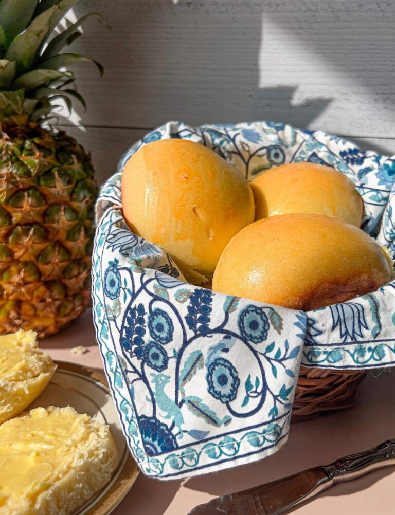 Sourdough Hawaiian rolls resting in a blue cloth lined basket with a pineapple in the background.
