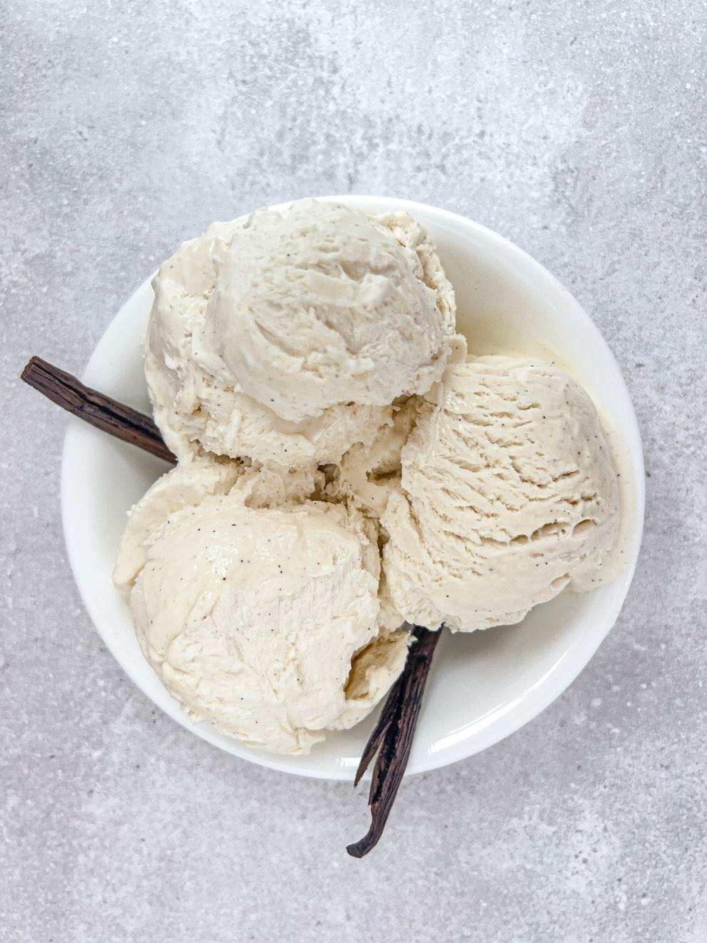 A bowl of three scoops of no churn vanilla bean ice cream in a white bowl on a grey background. 