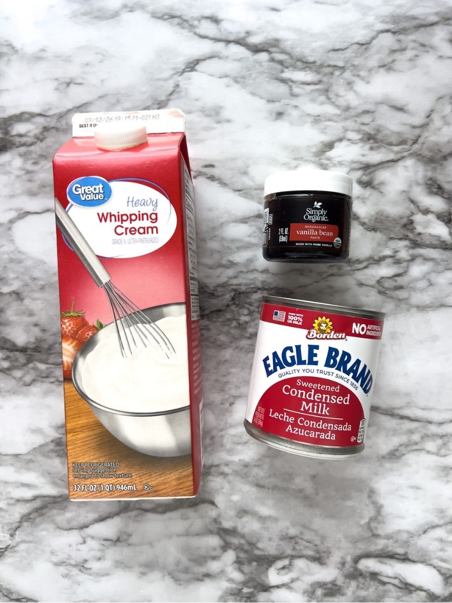 A carton of heavy whipping cream, a can of sweetened condensed milk, and a jar of vanilla bean paste on a marble background.