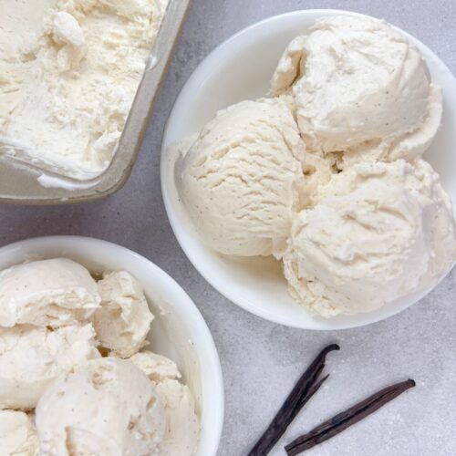 Two bowls of no churn vanilla bean ice cream on a grey textured background next to two vanilla bean pods.