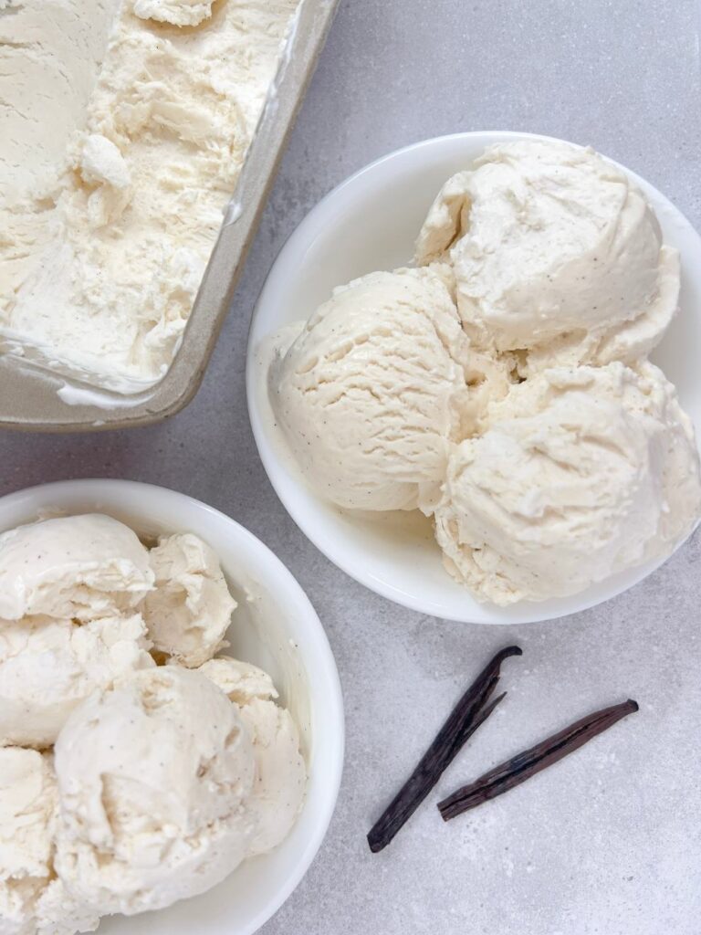 Two bowls of no churn vanilla bean ice cream on a grey textured background next to two vanilla bean pods.