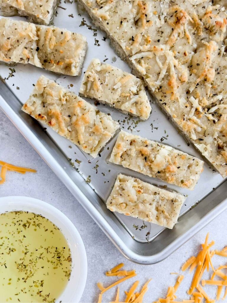 Cheddar Garlic Sourdough Focaccia Bread cut up into pieces on a baking sheet. A bowl of olive oil is in the bottom left corner.