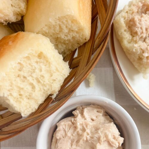 A basket of easy one day sourdough Texas roadhouse rolls next to a small bowl of honey cinnamon butter on top of a checkered tan and white tablecloth.