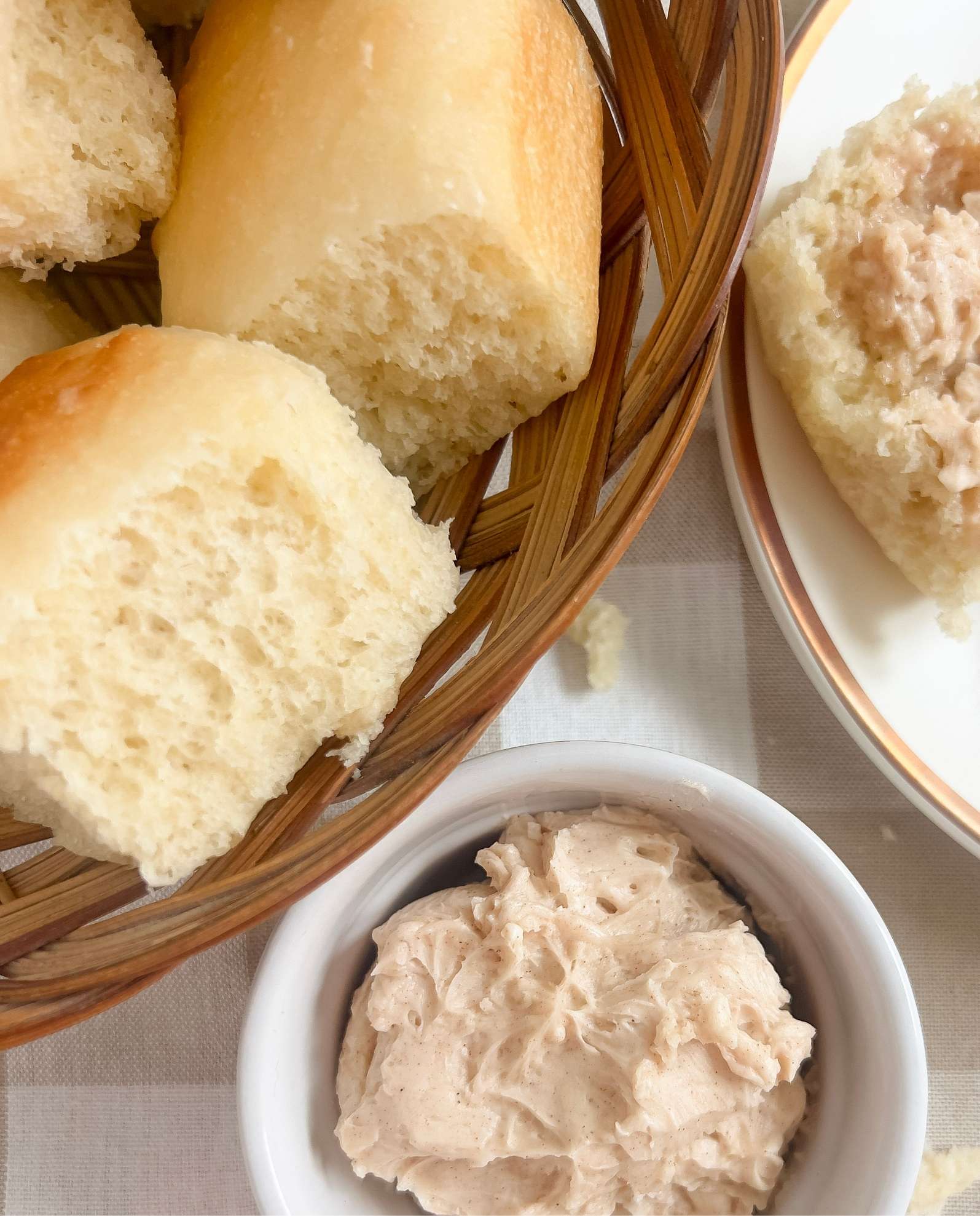 A basket of easy one day sourdough Texas roadhouse rolls next to a small bowl of honey cinnamon butter on top of a checkered tan and white tablecloth.