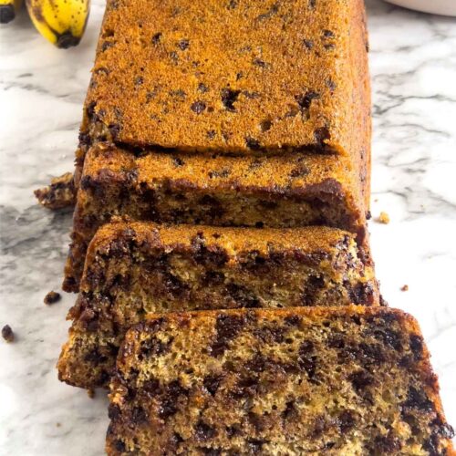 A loaf of loaded chocolate chip banana bread on a white marbled background with ripe bananas and chocolate chips in the background.