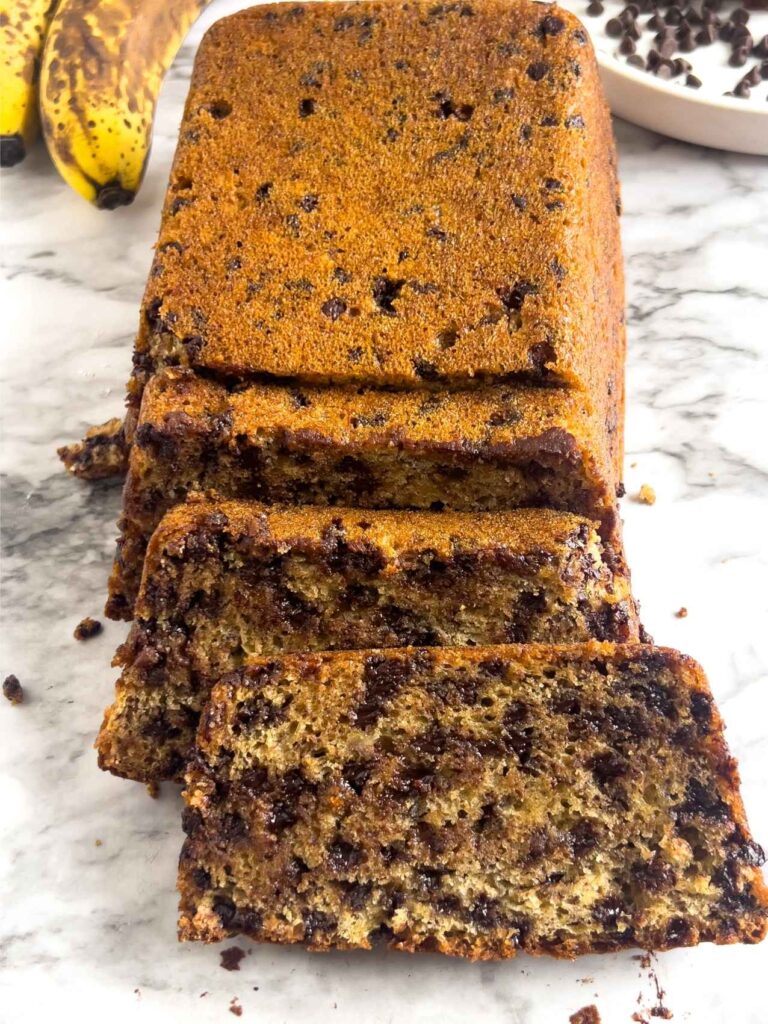 A loaf of loaded chocolate chip banana bread on a white marbled background with ripe bananas and chocolate chips in the background.