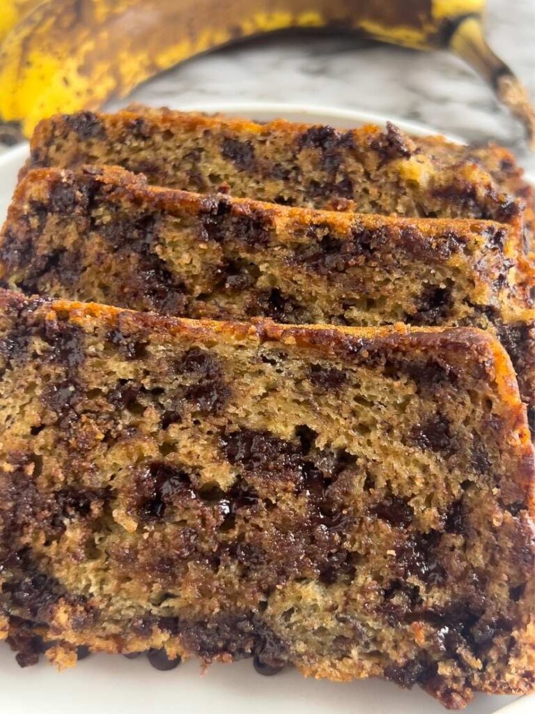 Slices of loaded chocolate chip banana bread on a marble background with a ripe banana in the background.