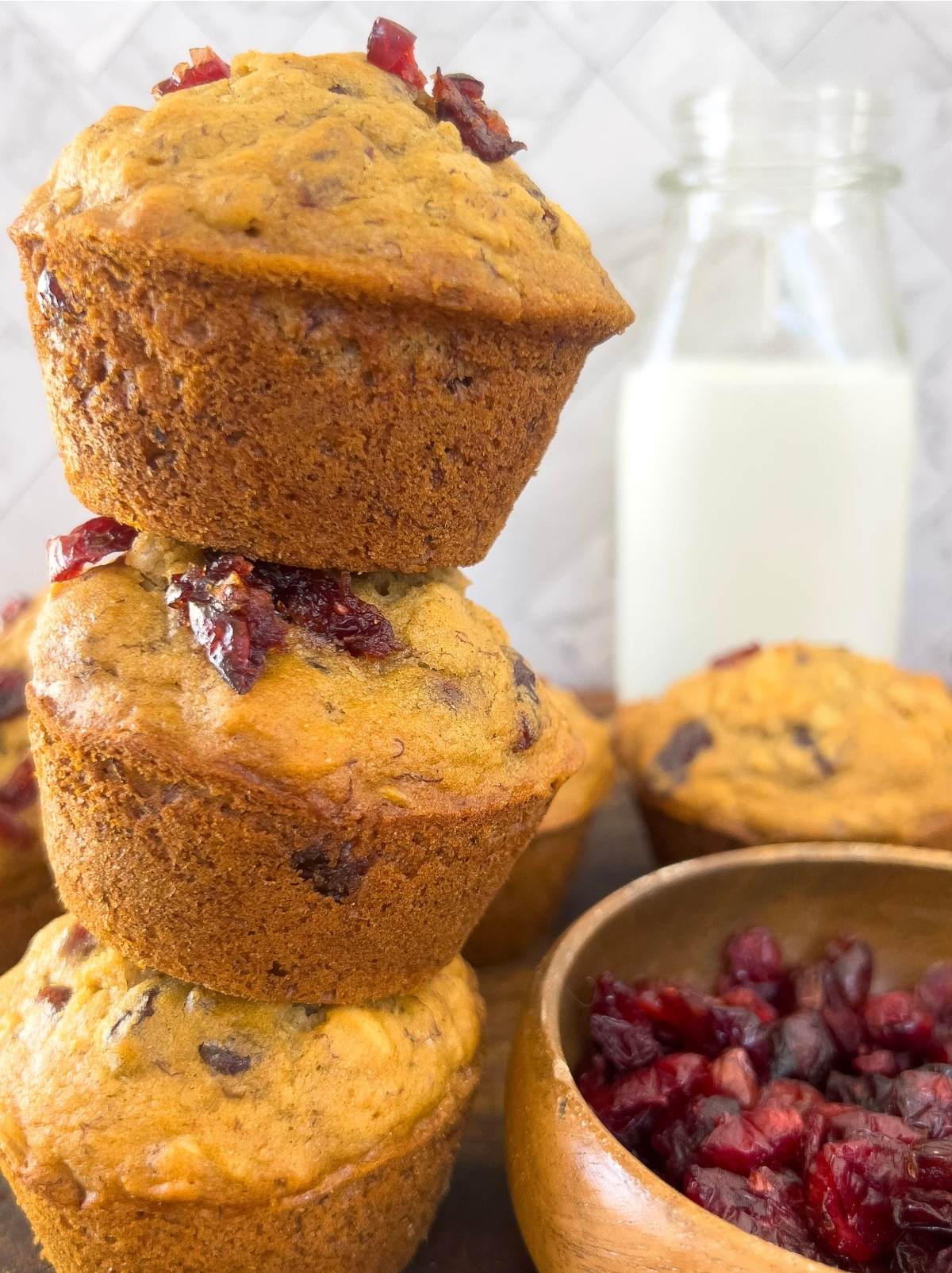 Three whole wheat cranberry banana oatmeal muffins stacked vertically in front of a marbled tile backsplash. A bowl of dried cranberries is nestled to the right of the muffins and a glass jug of milk is in the background.