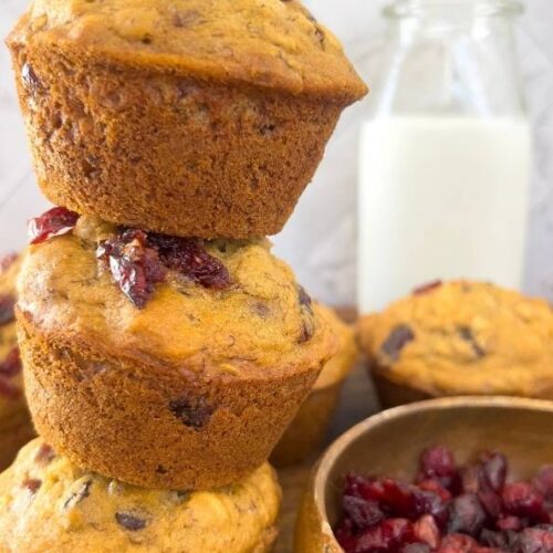 Three Whole Wheat Cranberry Banana Oatmeal Muffins stacked vertically with a dish of dried cranberries to the side and a jug of milk in the background.