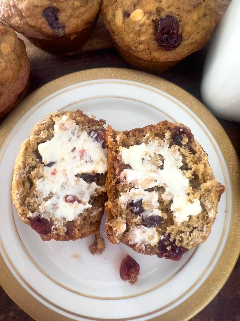 Whole Wheat Cranberry Banana Oatmeal Muffin split in half on a dish with a gold rim and covered in butter. There are three muffins in the top background along with a small jug of milk. 