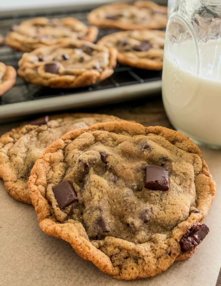 Two magnolia style chocolate chip cookies in front of a pan of cookies and next to a glass of milk.